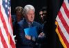 Bill Clinton standing between American flags with a folder.