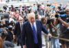 Donald Trump surrounded by media and bodyguards outside building.