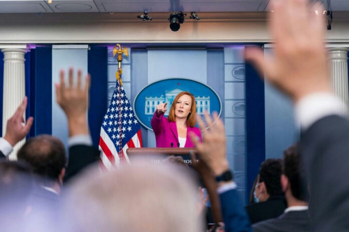 88d18a3b-59ed-4125-a391-b333f64f73a3-1024x683-1 Press secretary at podium, answering reporters questions, raised hands.