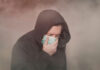 Person with hood and mask coughing in foggy setting.